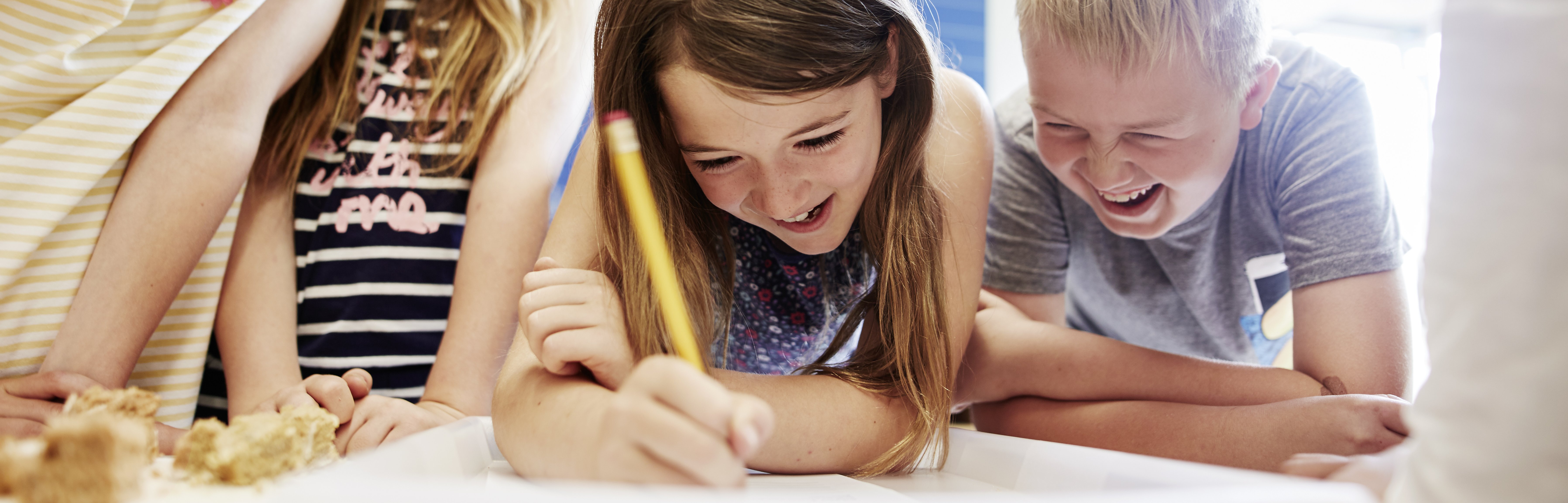 Kinder schauen gemeinsam auf ein Blatt Papier. Ein Mädchen schreibt gerade, ein Junge lacht.