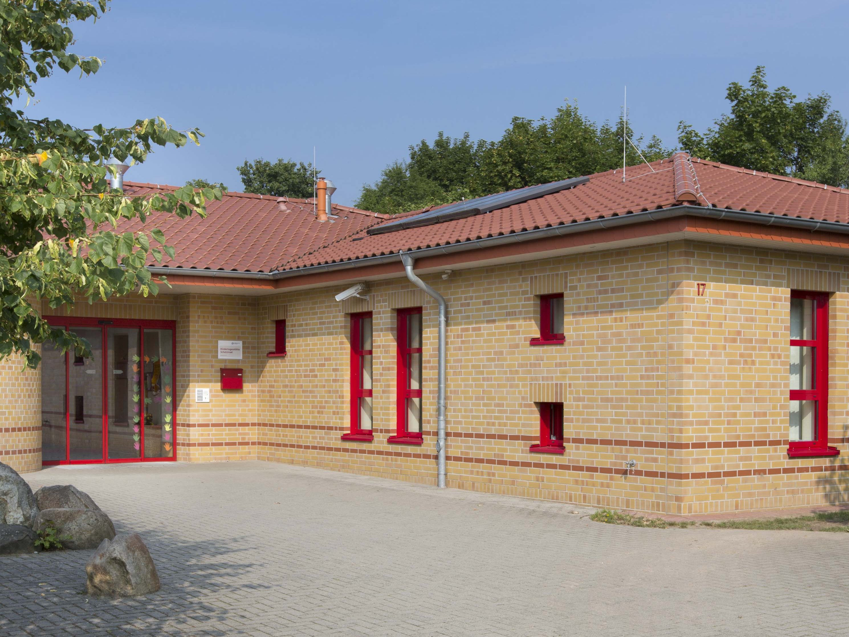 Das ist flache Gebäude ist aus sandbraunem Stein gebaut, die Fensterrahmen und Eingangstürrahmen sind Rot. 