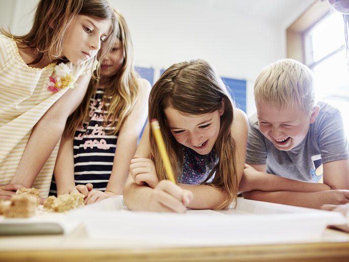 Kinder schauen gemeinsam auf ein Blatt Papier. Ein Mädchen schreibt gerade, ein Junge lacht.