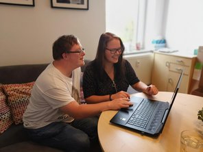 Ein junger Mann und eine junge Frau sitzen am Tisch und schauen gemeinsam auf ein aufgeklapptes Laptop. 