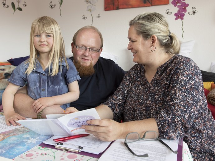 Ein Vater hat seine Tochter auf dem Schoss. Eine Mitarbeiterin berät die beiden.
