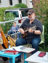 Mann sitzt auf grünem Stuhl und spielt Mandoline bei einem Terrassenkonzert, umgeben von Musikinstrumenten und Noten