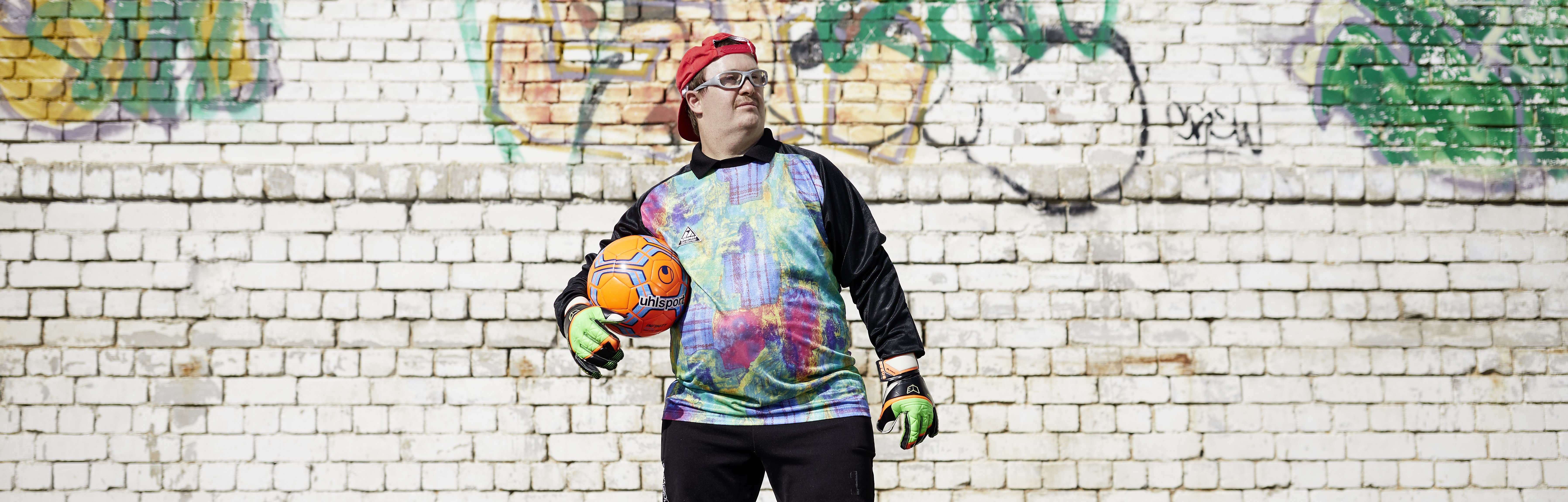 Ein junger Mann in sportlicher Kleidung steht vor einer Graffiti-Wand und hält einen Ball unter dem Arm.