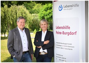 Uwe Hiltner und Susanne Speidel stehen draußen nebeneinander neben einer Stehle mit der Aufschrift Lebenshilfe Peine-Burgdorf und weiteren Kontaktinformationen. Im Hintergrund Natur - Bäume, Büsche, Wiese