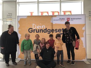 5 Kinder und zwei Erwachsene stehen vor einer Schauwand auf der steht "Dein Grundgesetz". Eine weitere erwachsene Frau sitzt im Schneidersitz vor der Gruppe.