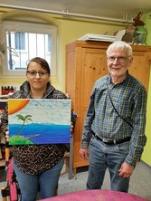 Auf dem Bild steht Frau L. neben dem Künstler Herr Carl. Frau L. zeigt ihr gemaltes Bild. Darauf ist eine Insel zu sehen, viel Meer und Himmel, die Sonne und ein Boot.