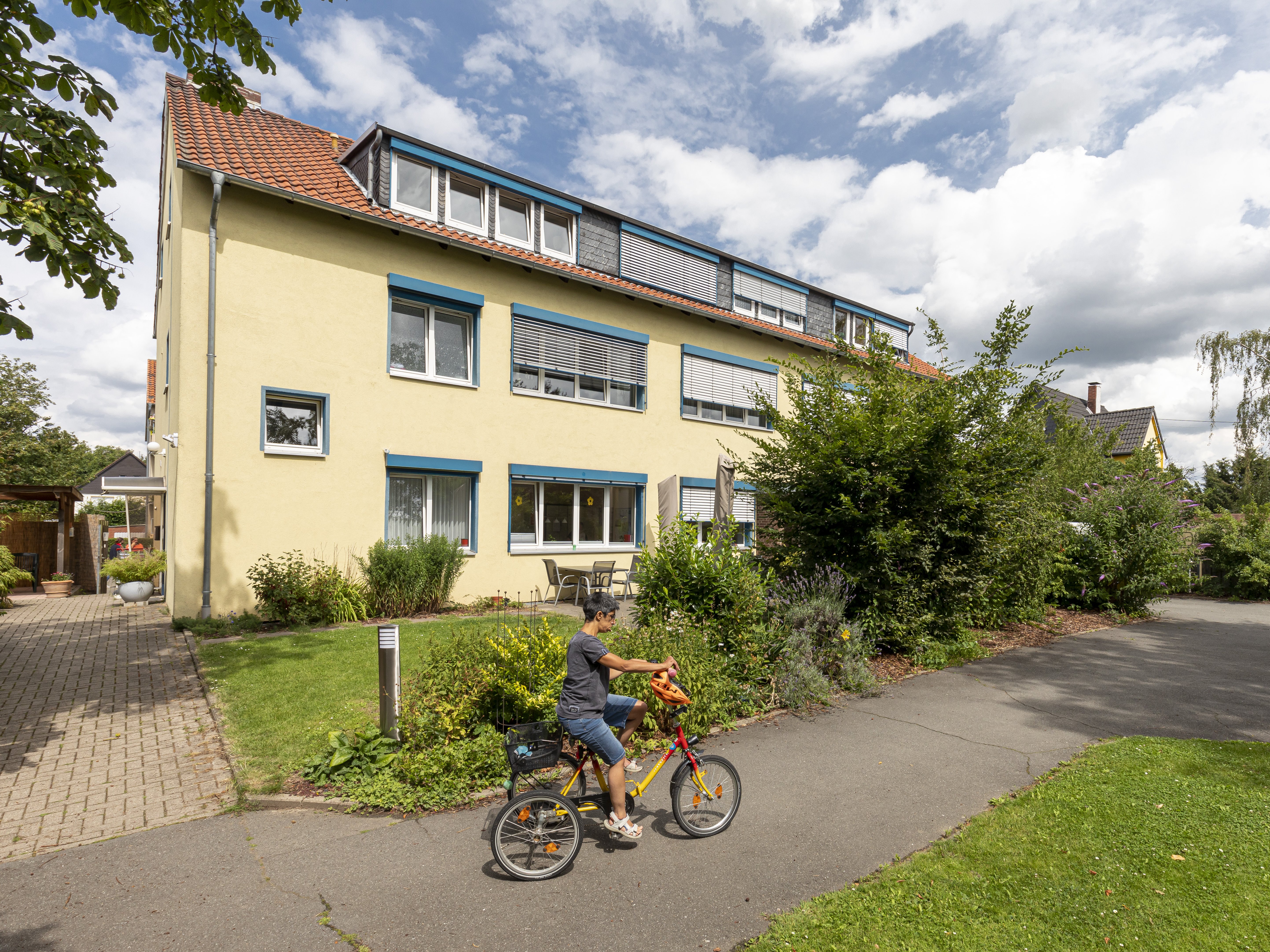 Haus hat drei Stockwerke, davon eines Dachgeschoss. Das Haus ist gelb und hat blaue Balken über den Fenstern.