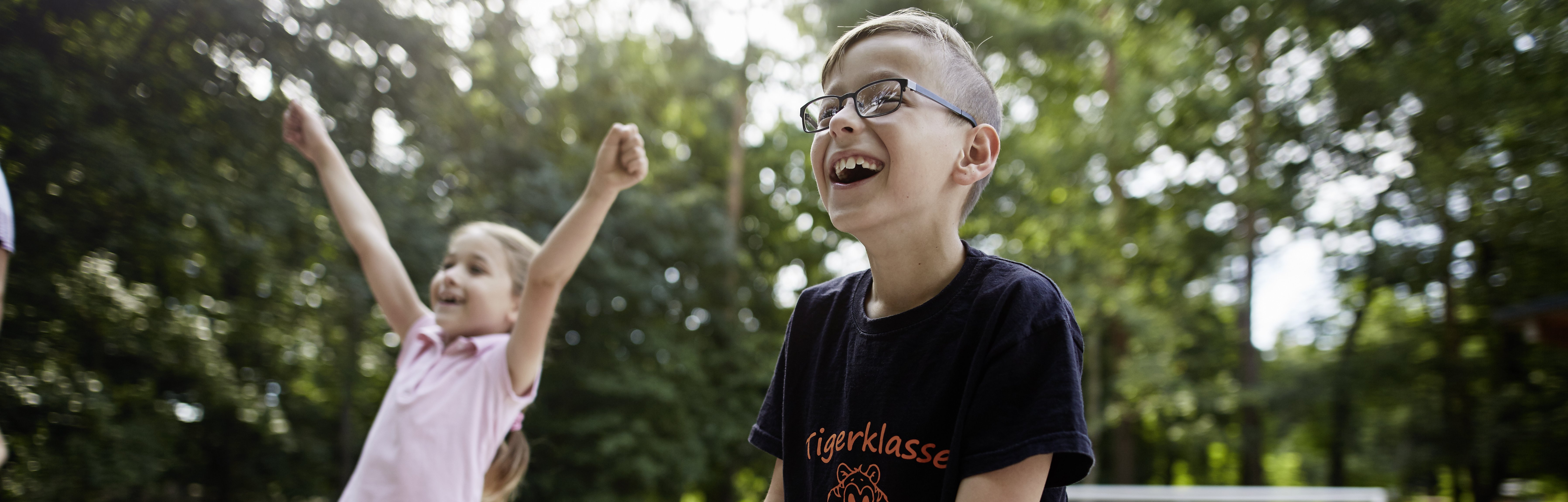 Zwei Kinder spielen lachen, eines reißt die Arme glücklich nach oben.