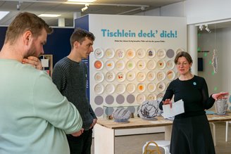 Das Bild zeiht eine Frau im Gespräch mit zwei Männern. Im Hintergrund sieht man gemalte Bilder, die ein Teil der Ausstellung sind.