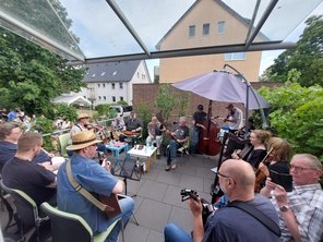 Musiker spielen bei einem Terrassenkonzert im Freien, Publikum sitzt auf Stühlen und genießt die Musik