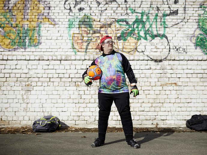 Ein junger Mann in sportlicher Kleidung steht vor einer Graffiti-Wand und hält einen Ball unter dem Arm.