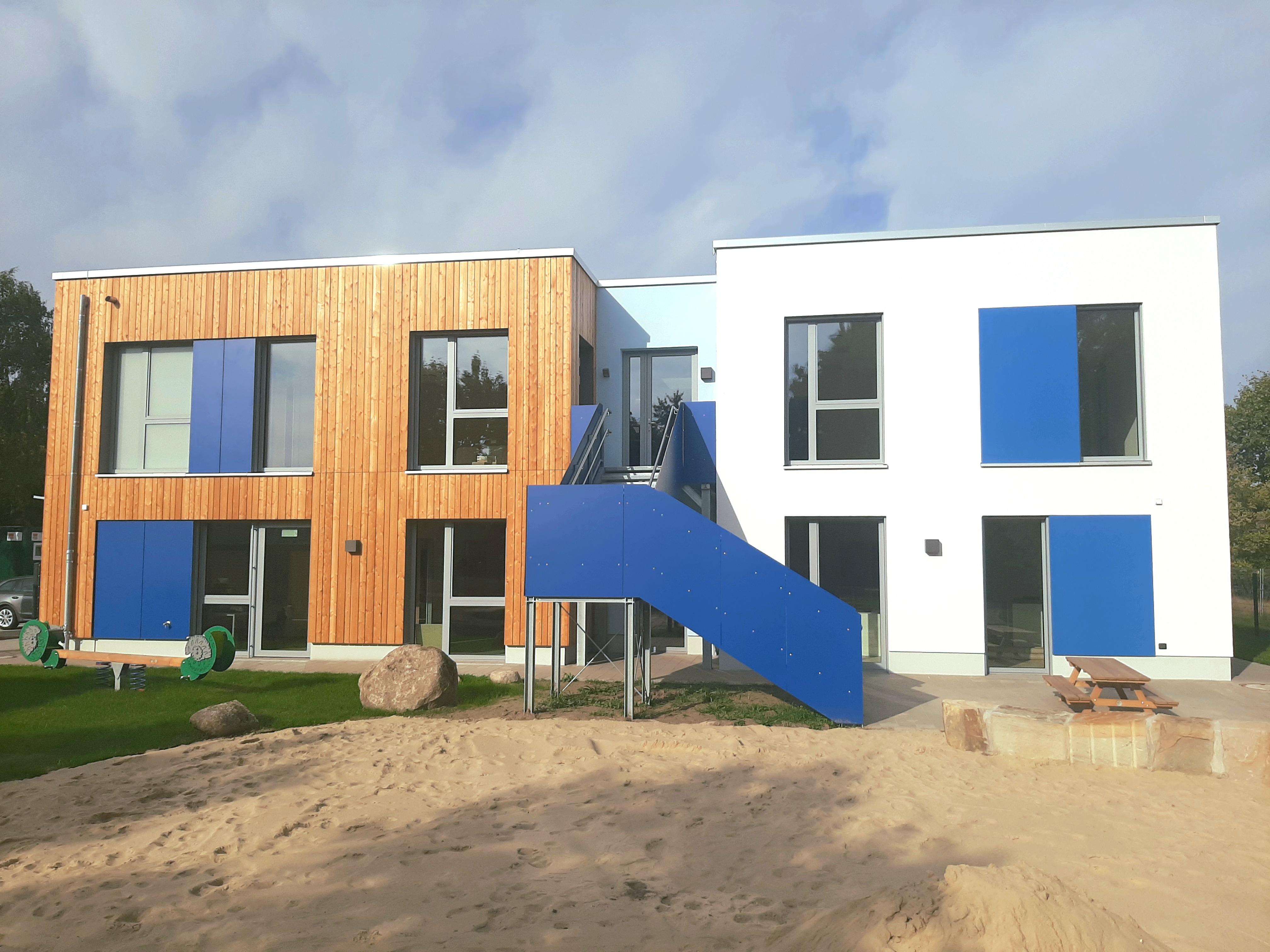 Seitenansicht des Gebäudes. Der linke Teil ist aus Holz, der rechte Teil ist weiß. Dazwischen ist eine blaue Außentreppe. Es gibt große Fenster, die Rahmen sind blau.